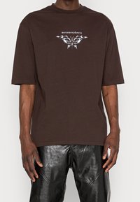 Brązowa, oversize'owa koszulka z bawełny, z graficznym wzorem zawierającym słowo "metamorfoza" w białym kolorze i motywami podobnymi do motyli.