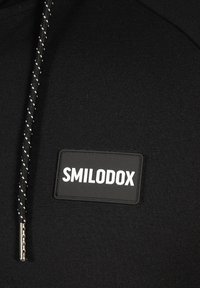 Sudadera negra con una tela texturizada. Presenta un distintivo de goma prominente con "SMILODOX" en blanco, y un cordón trenzado en blanco y negro.