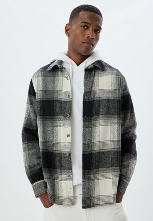 Geruite shirt in zwart en crème, gemaakt van structuurstof. Bevat knopen, een kraag en is gelaagd over een witte hoodie.