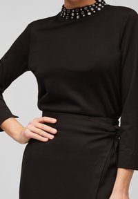 Czarny top z długim rękawem i wysokim kołnierzem, ozdobiony okrągłymi metalowymi ćwiekami, w połączeniu z czarną spódnicą typu wrap. Gładki materiał z dopasowanym krojem.