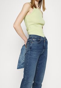 Persona vestida con una blusa sin mangas de color verde claro y jeans azules, sosteniendo un bolso de mano texturizado azul detrás de la espalda, contra un fondo blanco.