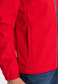 Giacca rossa con zip, polsini elastici e tasca laterale. Il tessuto appare liscio e resistente con un design aderente.