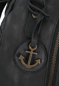 Detail van een zwarte leren tas met een ronde zwarte patch met een bronzen ankerembleem, en een zichtbare ritssluiting en stiksels.
