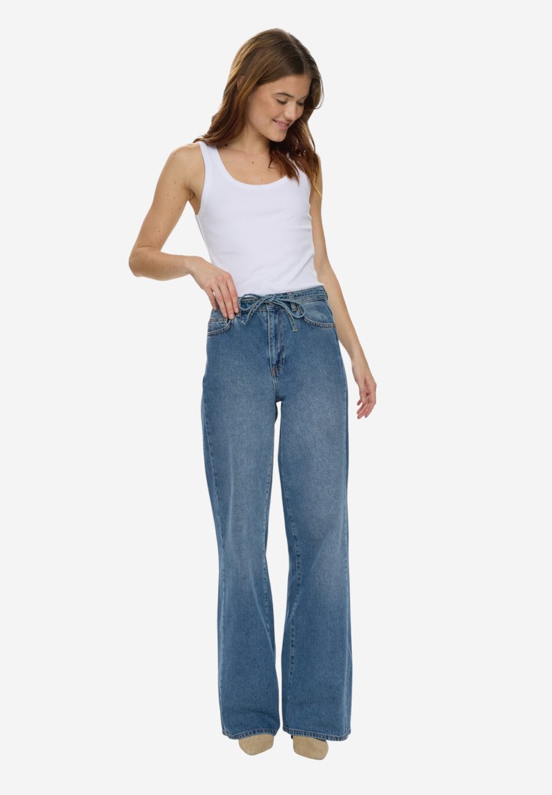 Femme portant un haut blanc sans manches et un jean bleu à taille haute et jambes larges, noué avec une ceinture à cordon, debout devant un fond blanc.