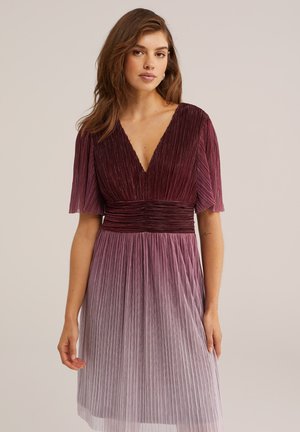 Žena v plisovaných šatách s krátkym rukávom a hlbokým výstrihom do tvaru písmena V, ktoré prechádzajú farbou zo tmavobordovej hornej časti do svetlomauve sukne.