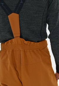 Bruine waterdichte broek met een elastische tailleband, verstelbare zwarte bretels en een gestructureerd oppervlak. Grijs languithemd eronder.