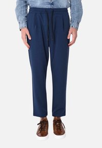 Pantaloni blu navy con pences, vita elastica e lacci neri. Abbinati a sneakers marroni con suole e lacci bianchi.