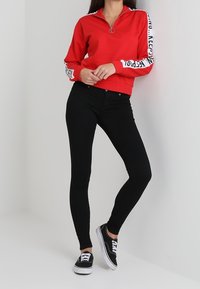 Sudadera roja con cremallera y detalles de logotipo blanco en las mangas, combinada con jeans skinny negros y zapatillas negras. Diseño ajustado, textura suave.