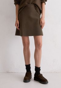 Vrouw die een bruine rok tot op de knie draagt, bijpassende trui, zwarte sokken en donkerbruine loafers, staand tegen een witte muur.