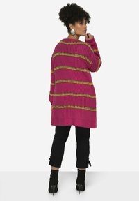 Lange roze gebreide cardigan met horizontale strepen in geel en groen, gedragen over zwarte broek met enkelbinders, gecombineerd met zwarte hakken.