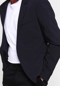 Man med en marinblå randig kavaj över en vit t-shirt och svarta byxor, med händerna i fickorna, visas från axlarna till höfterna.