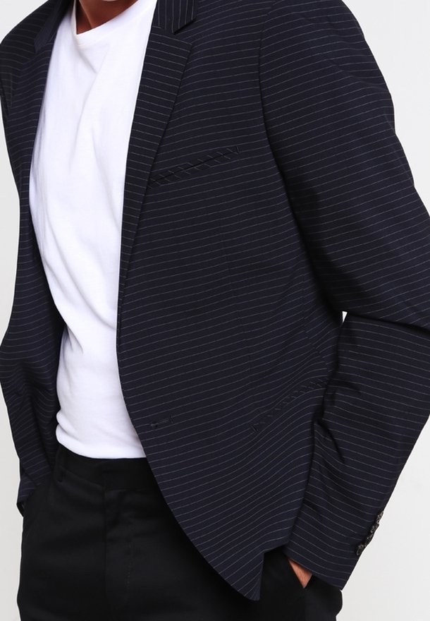 Man med en marinblå randig kavaj över en vit t-shirt och svarta byxor, med händerna i fickorna, visas från axlarna till höfterna.