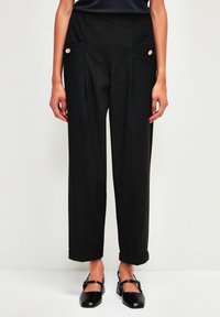 Korkeavyötäröiset, tummanharmaat housut, joissa on etupleatuja, sivutaskuja ja kultaisia nappikoristeita; käänteet; kankaassa on hieman rakennetta.