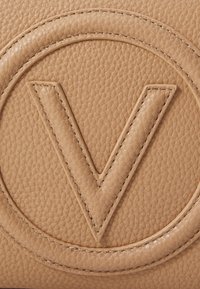 Sac en cuir texturé beige avec un design circulaire en relief présentant un "V" marqué et des coutures détaillées autour des bords.