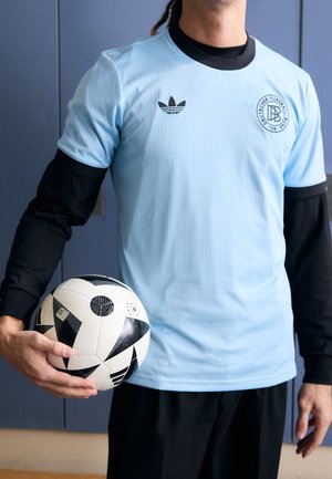 Persona con camiseta de fútbol azul clara sobre mangas largas negras sosteniendo un balón de fútbol negro y blanco contra una pared azul.