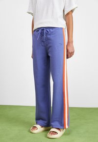 Paars trainingsbroek met oranje zijstrepen, een touwsluiting in de taille en een ontspannen pasvorm. G gedragen met lichtgekleurde sandalen op een groene ondergrond.