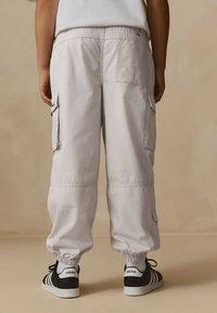 Pantaloni cargo gri deschis cu manșete elastice, dotate cu buzunare laterale și croială lejeră, asortați cu pantofi sport Adidas negri și albi.