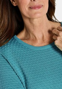 Türkiser Strickpullover mit strukturiertem Muster, Rundhalsausschnitt und kurzen Ärmeln, getragen von einer Person mit langen Haaren.