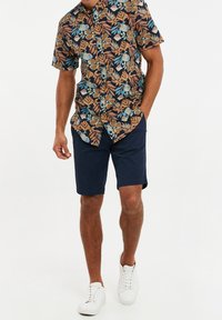 Korte mouwen, knoopsluiting shirt met een meerkleurig bloemen- en bladerenpatroon op een donkere achtergrond, gecombineerd met donkerblauwe shorts en witte sneakers.