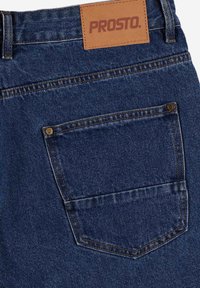 Granatowe dżinsy z ciemnego denimu z brązową skórzaną naszywką z napisem "PROSTO", z kontrastowym przeszyciem oraz tylną kieszenią z metalowym detalem w postaci nitki.