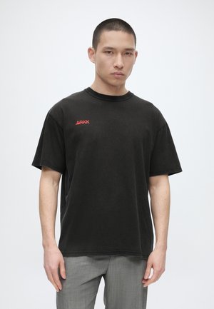 Ung mand med kort hår iført en sort ARKK t-shirt og grå bukser, stående foran en ensfarvet lys baggrund.