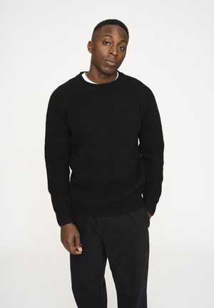 Sort ribbestrikket sweater med rund halsudskæring og lange ærmer, parret med mørke bukser, vist mod en hvid baggrund.