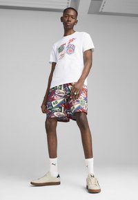 T-shirt bianca con stampe grafiche colorate, abbinata a pantaloni corti multicolore con vari loghi e motivi. Sneakers beige completano il look.