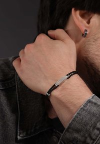 Bracelet tressé noir avec une plaque d'insigne de police en argent. Porté sur le poignet d'un homme, associé à une veste en denim gris.
