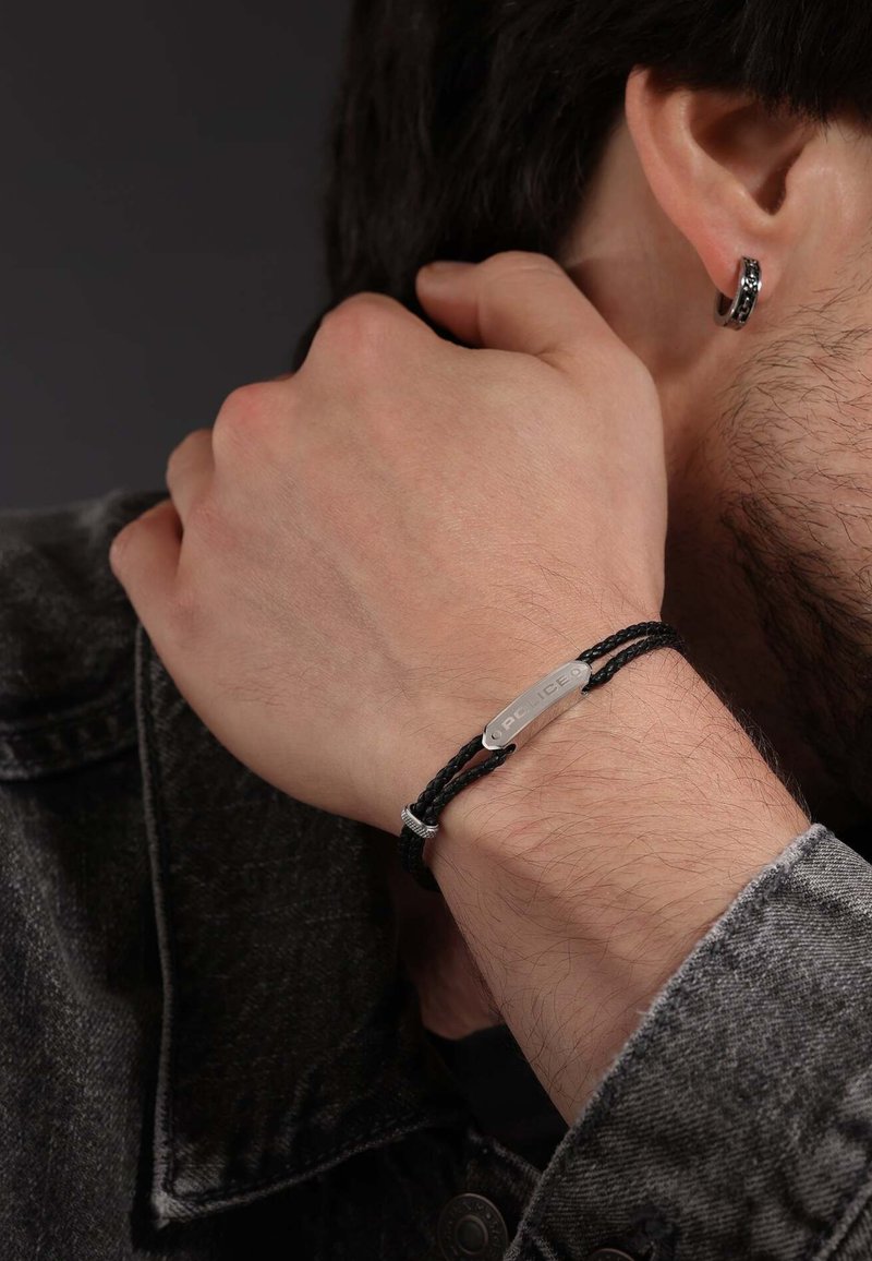 Bracelet tressé noir avec une plaque d'insigne de police en argent. Porté sur le poignet d'un homme, associé à une veste en denim gris.