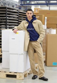 Hombre joven con abrigo y pantalones beige se apoya en cajas blancas apiladas sobre palés de madera dentro de un almacén con cajas de cartón en el fondo.