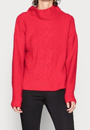 Femme portant un pull rouge vif en maille torsadée avec un col montant côtelé et des manches longues, associé à un pantalon noir.