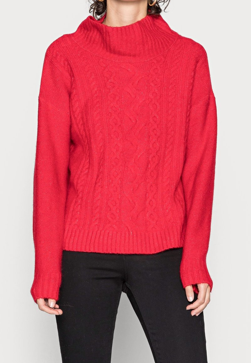 Femme portant un pull rouge vif en maille torsadée avec un col montant côtelé et des manches longues, associé à un pantalon noir.