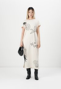 Donna che indossa un vestito maxi color crema con stampe botaniche nere, tiene in mano una borsa nera ed indossa stivali neri con tacco, in piedi contro un muro bianco.