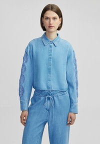 Camisa de denim azul claro con cuello y cierre de botones, que presenta intrincados detalles de ganchillo azul en las mangas.