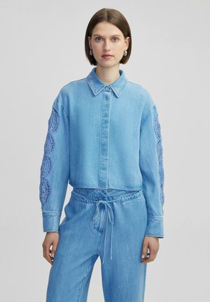 Light blue denim shirt with a collared neckline and button closure, featuring intricate blue crochet detailing on the sleeves.