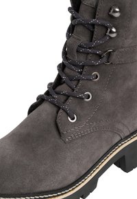 Botín de ante gris con cordones moteados en negro y blanco, ojales de metal, suela negra robusta y costuras blancas contrastantes.