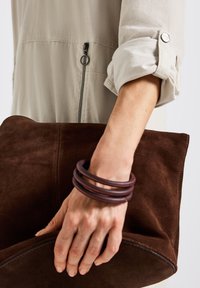 Käsi, joka pitää suurta tummanruskeaa mokkaisaldoa, päällä vaaleanbeige pitkähihaisempi paita, jonka hihat on nostettu ylös, ja tummat puukorut.