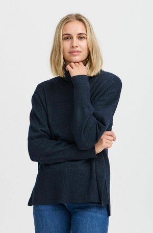 Pull bleu marine foncé avec un col haut, des poignets côtelés et des fentes sur les côtés, associé à un jean bleu, mettant en avant une texture de tricot douce.