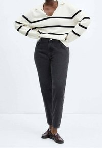 Suéter blanco con rayas negras, diseño de escote en V, combinado con jeans gris de cintura alta. Lleva unos mocasines oscuros y está de pie contra un fondo claro.
