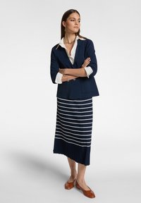 Blazer blu scuro sopra una camicia bianca con colletto, abbinato a una gonna a pieghe blu e bianca a righe. Le scarpe basse marroni completano l'outfit.
