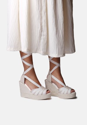 Weiße Keil-Espadrille-Sandalen mit Knöchelriemen, getragen zu einem weißen, strukturierten Midirock, die Füße und Unterschenkel vor weißem Hintergrund zeigen.