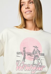 Sudadera de color crema con un gráfico de dos figuras sobre un círculo rosa, con la palabra "Wrangler" en rosa debajo. Material de mezcla de algodón.