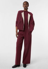 Bordowa kamizelka z wyściółką z sherpy, dopasowana bordowa bluzka i szerokie bordowe spodnie. Czarne botki do kostki dopełniają strój.