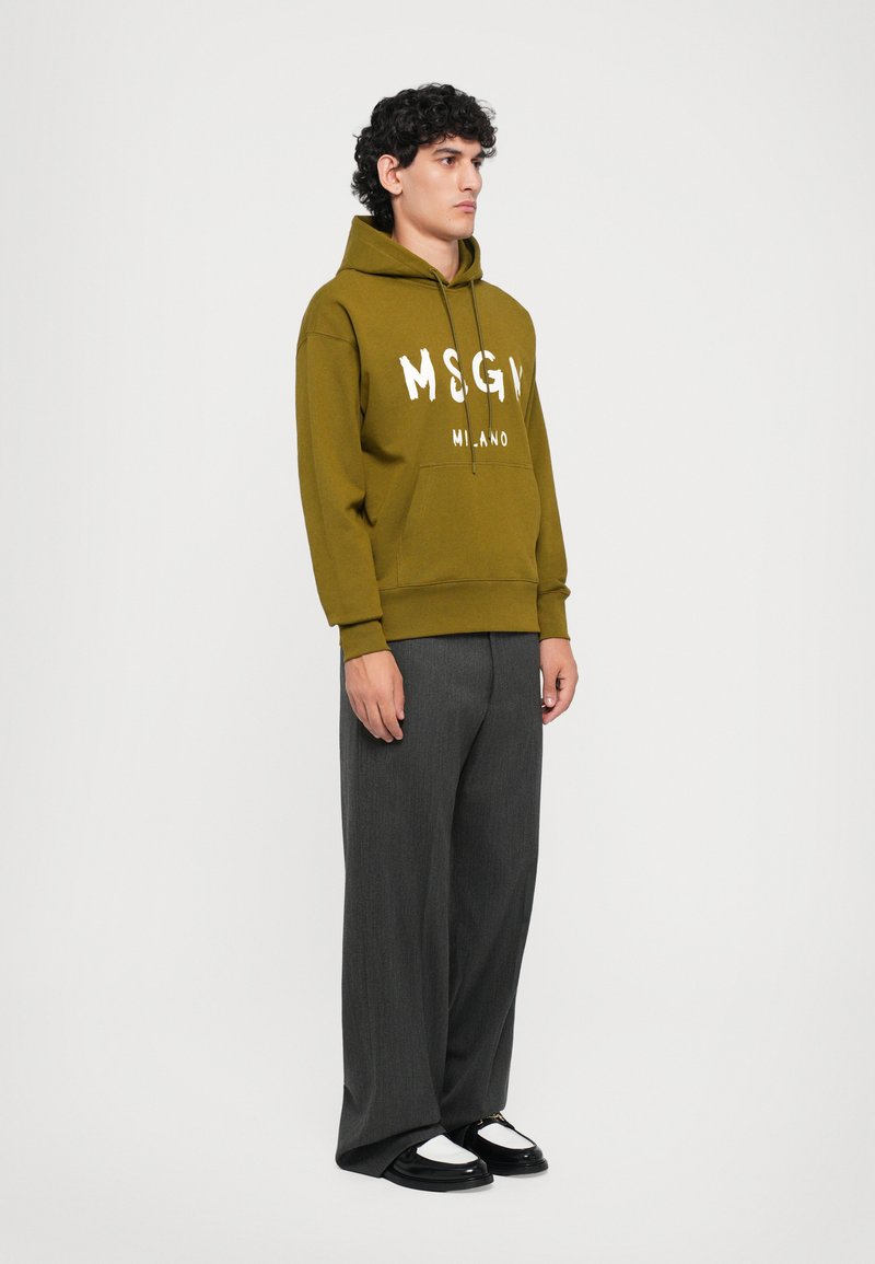 Homme se tenant debout portant un hoodie MSGM vert olive, un pantalon gris foncé ample et des mocassins noir et blanc, devant un fond clair uni.