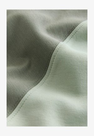 Minkšta medžiaga su žalia ir švelnia pilka spalvų blokų kompozicija. Lygi tekstūra, vientisa išlenkta kraštinė ir smulkios siūlės.