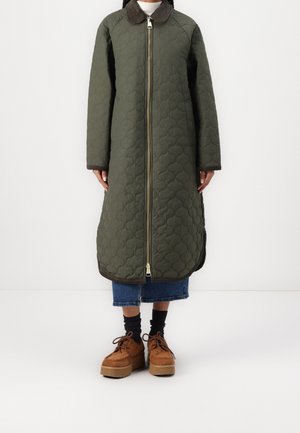 Osoba ubrana w długi, oliwkowozielony pikowany płaszcz z zamkiem z przodu, ciemny kołnierz, niebieskie spodnie dżinsowe o długości do kostek, czarne skarpetki i brązowe buty na platformie.