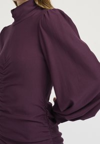 Vrchní díl švestkové barvy s vysokým límcem a naducanými rukávy. Má na bocích nabrání, které zdůrazňuje hladkou, pružnou texturu látky.