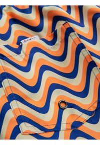 Tissu avec des rayures ondulées orange, bleues et crème, montrant une poche cousue avec un œillet métallique étiqueté "Ombre Wear."