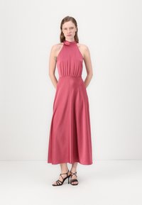 Roza halter haljina, do koljena, s lepršavom suknjom i pripijenim korzetom. Manekenka uparena s crnim sandalama na visoku petu. Glatka tekstura, minimalni detalji.