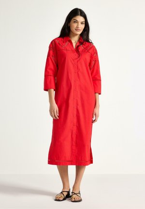 Kvinde iført en lang, rød kjole med knapper foran, 3/4-ærmer og sorte sandaler med stropper, stående mod en ensartet lys baggrund.